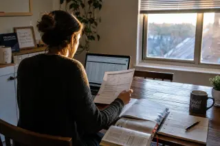 Early morning home office with sunlight on a wooden table, accounting books, notes, coffee mug, and a focused young entrepreneur working from behind.
