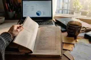 Close-up of a hand turning pages of a French accounting ledger on a busy home office desk, with paperwork, a coffee mug, soft laptop glow, and warm morning light.